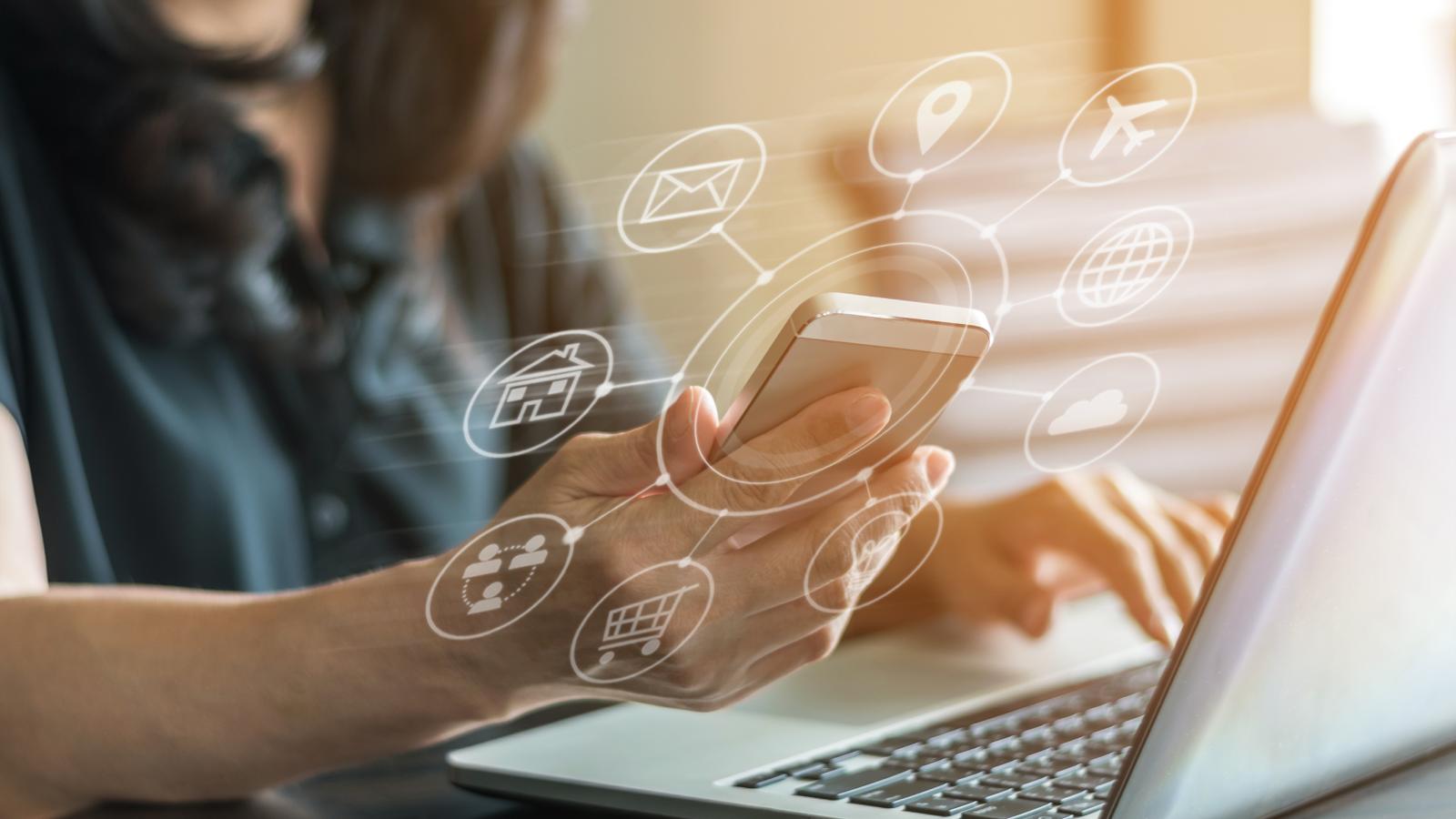 Lady sat at laptop, holding a mobile phone which is surrounded by icons